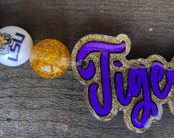 May include: A purple and gold beaded lanyard with a purple "Tigers" charm. The charm has a gold glitter outline and purple lettering. The lanyard also features beads in purple, white with an LSU logo, and gold glitter.