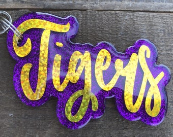 May include: A purple glitter keychain with the word "Tigers" in gold script. The keychain has a silver clasp and a ring for attaching keys. The design is layered, creating a 3D effect. The background is a weathered wooden surface.