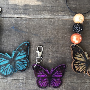 May include: A collection of butterfly-shaped accessories, including keychains and a necklace. The keychains feature blue and purple glitter butterflies with silver clasps and beaded accents. The necklace has a gold glitter butterfly pendant and orange, black, and gold beads.