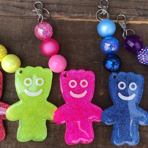 May include: A collection of colorful keychains featuring glittery Sour Patch Kids-shaped charms in various colors, including red, green, pink, blue, and purple. Each charm is attached to a beaded strand with a silver clasp.