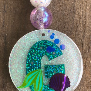 May include: A round, glittery keychain with a teal letter "G" and mermaid tail design. The keychain has a purple seashell and blue bubbles. The keychain is attached to a beaded strand with turquoise, white, and purple beads.