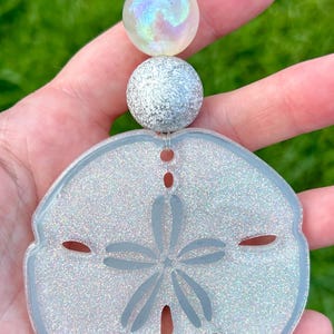May include: A shimmering, light blue sand dollar ornament with a glittery finish, featuring a star-shaped design in the center. The ornament is connected to three decorative, glittery beads and a silver wire loop for hanging.