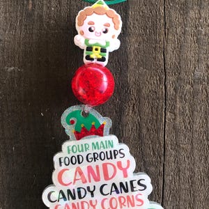 May include: A Christmas-themed keychain with a green wire loop. It features a cartoon elf, a red glittery ball, and a tree-shaped charm with the text "FOUR MAIN FOOD GROUPS CANDY CANDY CANES CANDY CORNS & SYRUP."