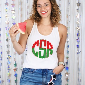 May include: White tank top with a red and green watermelon monogram. The monogram is in a circle with a scalloped edge. The person is holding a slice of watermelon.