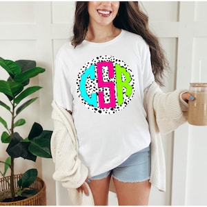 May include: White t-shirt with a black and white polka dot circle design featuring a monogram with the letters "C", "S", and "R" in bright pink, turquoise, and green.