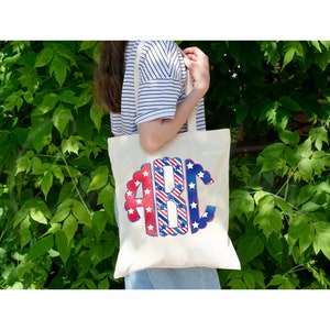 K&ouml;nnte beinhalten: Eine wei&szlig;e Canvas-Tragetasche mit einem rot-wei&szlig;-blauen Monogramm-Design mit den Buchstaben "ABC". Das Monogramm ist von einem gewellten Rand umgeben. Die Tasche wird von einer Person getragen, die ein gestreiftes Hemd und eine blaue Jeans tr&auml;gt.