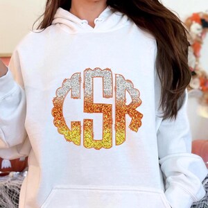 May include: White hoodie featuring a large, glittery candy corn monogram. The monogram is in a circle with a scalloped edge and displays the letters "CSR" in orange, yellow, and white.
