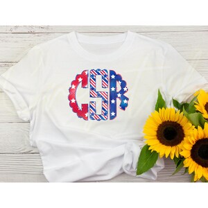 K&ouml;nnte beinhalten: Wei&szlig;es T-Shirt mit einem rot-wei&szlig;-blauen Monogramm-Design. Das Monogramm hat die Form eines Kreises mit Sternen und Streifen. Das Shirt liegt auf einem Holzgrund.