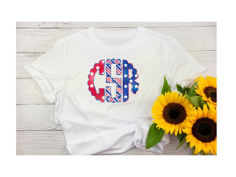 K&ouml;nnte beinhalten: Wei&szlig;es T-Shirt mit einem rot-wei&szlig;-blauen Monogramm-Design. Das Monogramm hat die Form eines Kreises mit Sternen und Streifen. Das Shirt liegt auf einem Holzgrund.