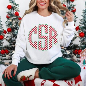 May include: White sweatshirt with a red and white candy cane striped monogram. The monogram is in a circle with a green outline. The sweatshirt is worn by a person sitting on a white and red blanket.
