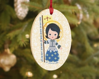 May include: Oval wooden Christmas ornament featuring a cartoon illustration of Saint Joan of Arc in a blue and gold dress holding a banner with a cross. The ornament has the text "I am not afraid I was born to do this - St. Joan of Arc" printed on it.