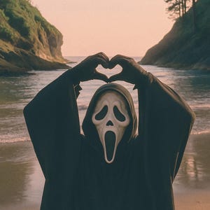 May include: A person in a black hooded robe and a Scream mask forms a heart shape with their hands against a beach backdrop. The mask is white with a gaping mouth and hollow eyes. The scene is set at sunset.