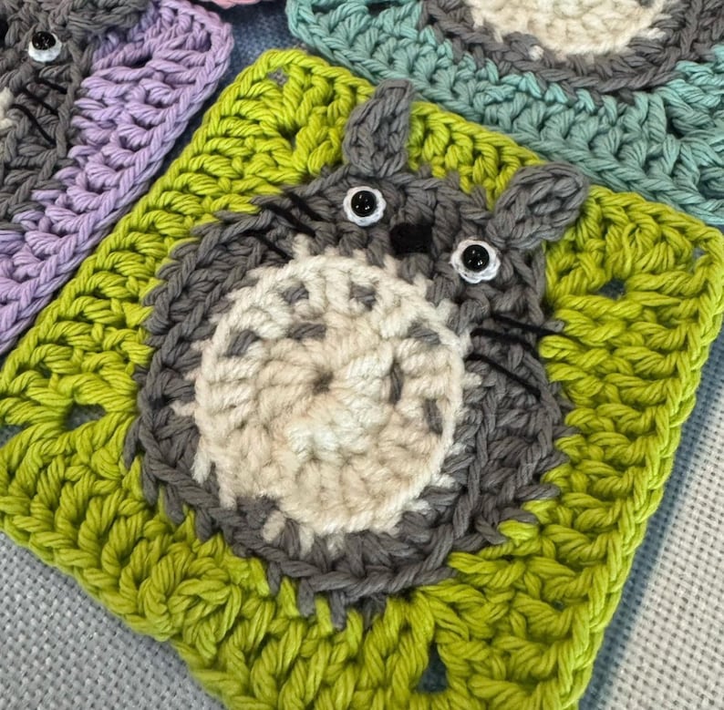 Pu&ograve; includere: Un quadrato all'uncinetto verde con un personaggio di Totoro all'uncinetto grigio al centro. Il Totoro ha una pelliccia all'uncinetto bianca e occhi all'uncinetto neri.