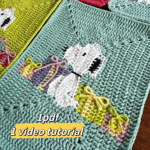 May include: A crochet square featuring a white dog with black ears and a green, yellow and pink gift box. The square is made with a light green yarn and has the text "1 pdf 1 video tutorial" at the bottom.