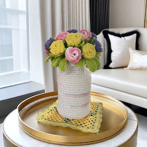 May include: A crocheted bouquet of yellow, pink, and purple flowers in a white crocheted vase. The vase is sitting on a gold tray with a green crocheted doily.
