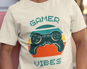 May include: A person wearing a beige t-shirt with a retro-style graphic of a video game controller and the text "GAMER VIBES".