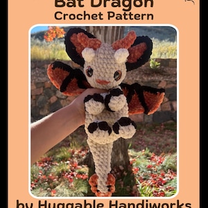 May include: A crocheted bat dragon toy in cream, orange, and black. The toy has large wings, a long tail, and is labeled "Ember the Bat Dragon Crochet Pattern" by Huggable Handiworks. The background shows a tree and foliage.