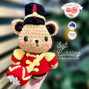May include: A crocheted plush toy bear wearing a red and yellow uniform with a black hat. The bear is holding a red and yellow drum. The text "Happy Hug" is in the top right corner of the image. The text "Soft Cuddling" is in the bottom centre of the image. The text "Famcraftshop - Lucky Crochet Mates" is below the text "Soft Cuddling".