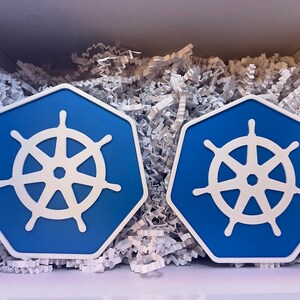 May include: Two blue and white hexagonal coasters with a ship's wheel design. The coasters are set against a white background with shredded paper, suggesting a gift box presentation. The coasters are a vibrant blue with a white wheel.