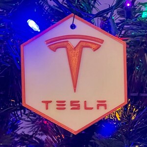 May include: A white and orange hexagonal ornament with the word "TESLA" printed in orange. The ornament features the Tesla logo in orange. The ornament is hanging on a Christmas tree with blue and green lights.