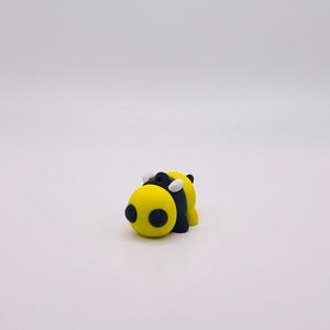May include: A small, yellow and black bee figurine with a rounded body and large, black eyes. The bee has white accents on its back and is set against a plain white background. This handcrafted item is a charming decorative piece.