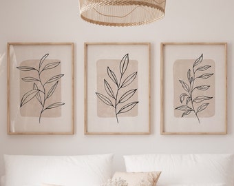 May include: Three framed prints with minimalist line drawings of leafy branches on a beige background. The prints are hanging on a white wall above a white sofa with a beige throw blanket and a woven coffee table.