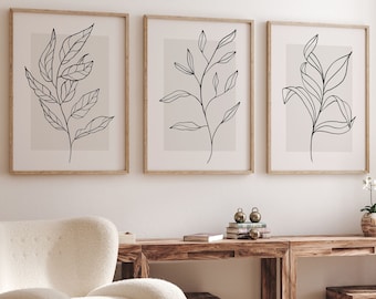 Puede incluir: Tres impresiones minimalistas de arte lineal de ramas frondosas en negro sobre fondo blanco, enmarcadas en madera natural, colgadas en una pared blanca sobre una mesa de madera y un sillón blanco.