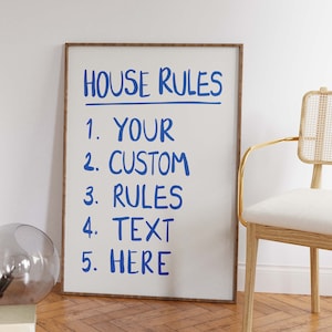 Op de afbeelding: Een witte poster in een lijst met de tekst "HOUSE RULES" in blauwe handgeschreven letters. De poster somt vijf regels op, met de nummers 1 tot en met 5, in blauwe handgeschreven letters. De regels zijn "YOUR", "CUSTOM", "RULES", "TEXT" en "HERE". De poster hangt aan een witte muur naast een licht houten stoel met een wit kussen.