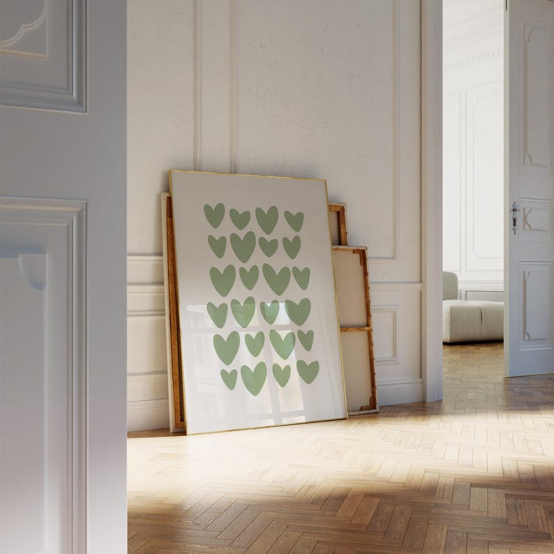 May include: A framed art print featuring a pattern of sage green hearts on a white background. The artwork is displayed in a room with a wooden floor and white walls, leaning against other framed pieces.