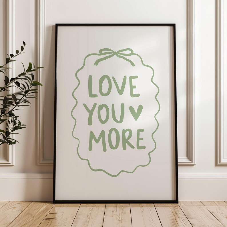 May include: A framed print with the words "LOVE YOU MORE" in green script. The text is inside a green, wavy border with a bow at the top. The frame is black, and the background is white. The print is displayed on a wooden floor.