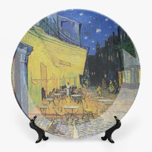 May include: Decorative plate featuring a painting of a nighttime cafe scene with yellow walls, tables, and chairs. The artwork depicts people dining under a starry sky. The plate is displayed on a black stand.