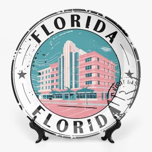 Op de afbeelding: Een decoratieve witte plaat met een roze en blauwe illustratie van een gebouw en de tekst "FLORIDA" in een cirkelvormig stempelontwerp. De plaat wordt gepresenteerd op een zwarte standaard.