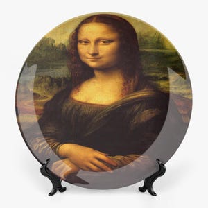 Op de afbeelding: Een decoratieve plaat met het schilderij van de Mona Lisa. De plaat heeft een glanzende afwerking en wordt gepresenteerd op een zwarte standaard. Het kunstwerk toont een vrouw met een subtiele glimlach, gekleed in een donkere jurk, met een landschapsachtergrond.