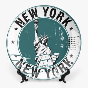 Op de afbeelding: Witte keramische plaat met het Vrijheidsbeeld in een turquoise cirkel, met de woorden "NEW YORK" in een cirkelvormig patroon. De plaat heeft een vintage, distressed ontwerp met ster- en stempeldruk.