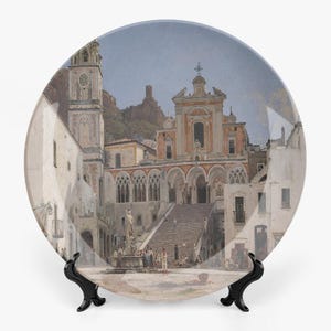 Op de afbeelding: Decoratieve plaat met een gedetailleerd schilderij van een Europees stadsplein met een kerk, fontein en mensen. De plaat wordt gepresenteerd op een zwarte standaard. Het kunstwerk heeft een vintage esthetiek met gedempte kleuren en architecturale details.