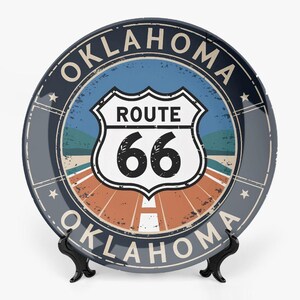 Op de afbeelding: Een decoratieve plaat met een Route 66-bord met het nummer 66 in het midden. De plaat heeft een blauw en oranje kleurenschema met het woord "Oklahoma" in een cirkelvormig patroon rond de rand. De plaat wordt op een zwarte standaard gepresenteerd.