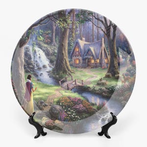 May include: A decorative plate depicting a forest scene with a cottage, waterfall, and a figure in a yellow dress. The plate is supported by a black stand. The artwork has a fairytale theme with vibrant colours and detailed imagery.