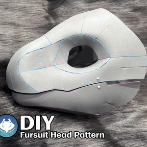 Peut inclure: Une base de tête de fursuit en mousse gris clair présentant une forme animale sculptée avec une grande ouverture pour l'œil, marquée de lignes à l'encre bleue pour l'assemblage et deux petites épingles blanches. Le texte DIY Fursuit Head Pattern est visible.