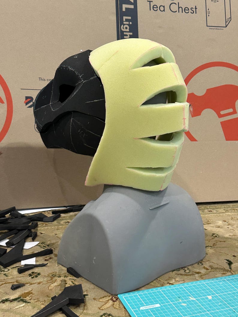 May include: A black and green foam mask with cutouts for eyes and mouth, being fitted on a gray mannequin head. The mask is being constructed with foam pieces.