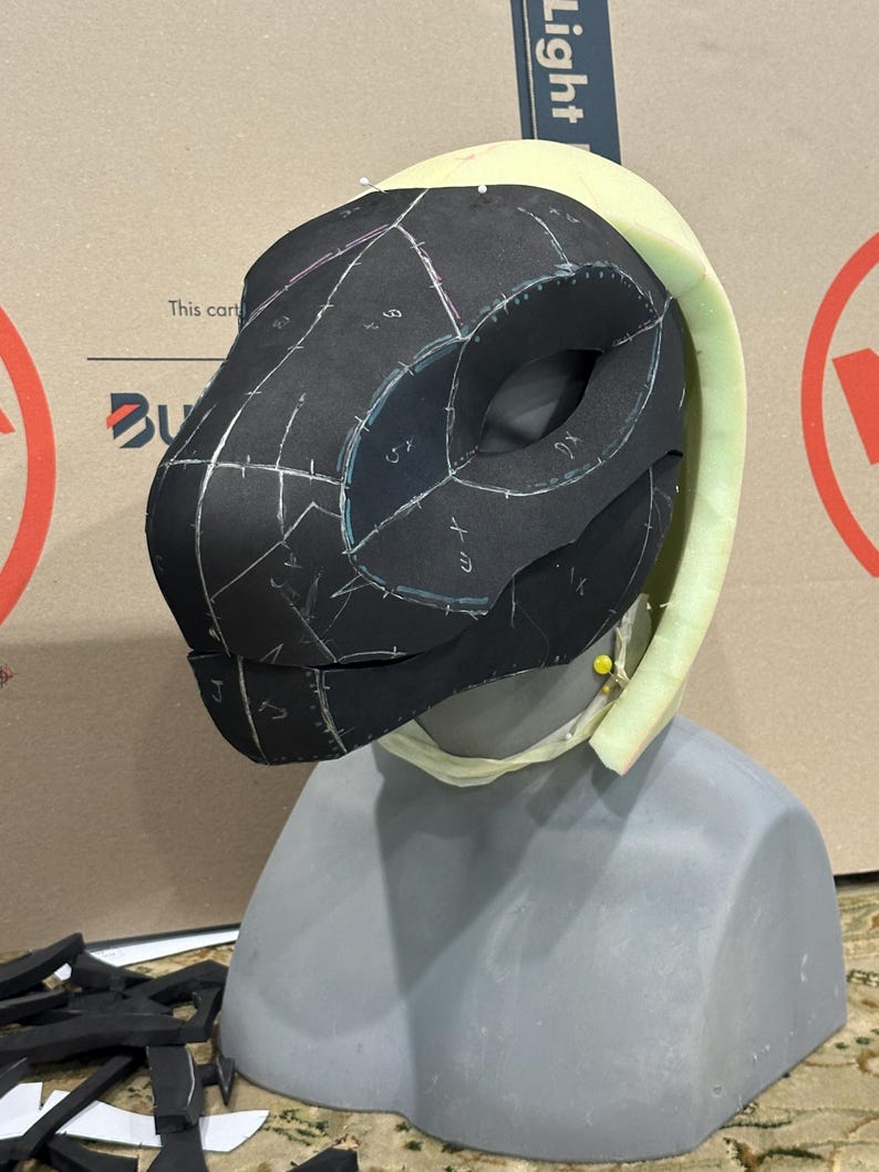 May include: A black foam mask with white markings and numbers, pinned to a white foam head form. The mask is being prepared for a costume or cosplay project.
