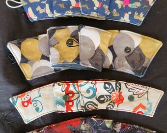 May include: A set of 10 colorful fabric coffee cup sleeves. The sleeves are made of different fabrics and feature various patterns, including animals, flowers, and geometric designs. Each sleeve has a button closure.