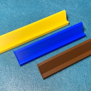 May include: Three L-shaped display stands arranged diagonally. One stand is bright yellow, one is vibrant blue, and one is a solid brown. Each stand features a flat base and a vertical back panel, designed to hold items upright in a stable position.