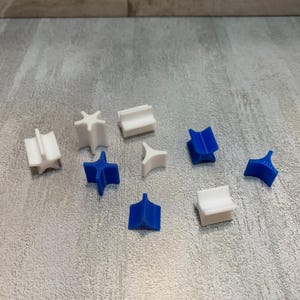 May include: A collection of small, geometric 3D-printed objects in white and blue. The shapes include star-like forms, curved pieces, and block-like designs. The objects are scattered on a textured, gray surface.