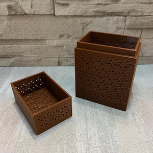 May include: Two-piece brown decorative storage box set. The larger box is rectangular with a removable lid. Both pieces feature a repeating pattern of small, leaf-shaped cutouts. The boxes are made of a solid, matte material.