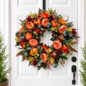 May include: A circular wreath featuring orange pumpkins, vibrant orange and red roses, green eucalyptus leaves, yellow filler flowers, and dark red spiky accents. The arrangement is dense and multi-textured, displaying a variety of autumn-inspired colors.