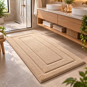 May include: A rectangular beige bath mat with a textured, shaggy pile surface and a distinct raised border frame design. The mat features a soft, plush appearance and is positioned flat on a tiled floor in a bathroom setting with neutral tones.