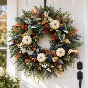 May include: A circular autumn wreath featuring white pumpkins, dark blue flowers, and clusters of white and red berries. The arrangement includes green eucalyptus leaves, sage-colored foliage, and orange-toned leaves on a round base.