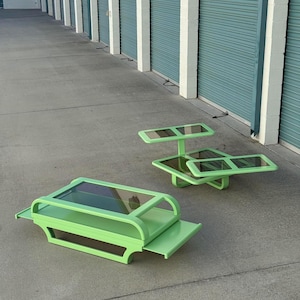 May include: Two green, retro-style coffee tables with glass tops. One is rectangular with a lower shelf, and the other is a tiered, asymmetrical design. The tables are painted in a bright, solid green color.