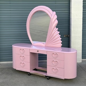 May include: A pastel pink vanity table featuring a large oval mirror with a fan-shaped side accent. The base includes six drawers, each adorned with a heart-shaped pull. The entire piece is finished in a smooth, glossy pink paint and is mounted on small wheels.