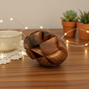 May include: A wooden puzzle ball with a geometric design. The ball is made of interlocking wooden pieces in shades of brown. The puzzle sits on a wooden surface, with a small bowl and string lights in the background.